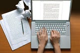 A pair of a woman’s hands typing on a laptop keyboard with the screen showing a full written page. At the left side of the laptop,  several pieces of typed paper and a pencil rest on the table, with three crumpled pieces scattered on the top side of the papers.