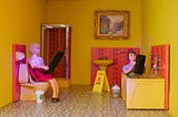 Two dolls in a tiny pink and yellow bathroom, one on the toilet and one in the bathtub, both looking at giant smartphones.