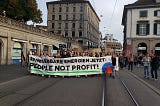 Ein Fronttransparent — Erneuerbare Energien Jetzt! People not Profit mit dem Klimastreiksymbol. Demozug dahinter auf einer Strasse in Zürich