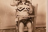 Old black & white photo of little boy sitting in a chair and holding a ball.