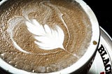 Close up of a flat white coffee with latte art in the shape of a feather, on a “Black and White” branded cup and plate.