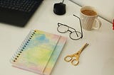Photo of a computer keyboard and mouse, spiral bound journal, scissors, glasses, cup of coffee.