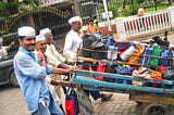 Four Indian men, all wearing white cloth caps push trailers loaded with lunch tins and bags.