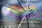 rainbow Pride flag waving on a flagpole over a building, seen through glass shattered by a bullet