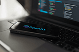 OpenAI logo glowing in blue-purple on smartphone screen placed on laptop keyboard with code editor in background.