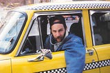 Bearded cab driver with baseball hat on backwards leaning out the window of an old time Checker taxicab