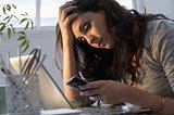 A millennial woman who feels depressed sits at her desk, clutching her forehead and looking at her phone.