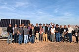 A group of people smiling and standing together in front of 3 solar panels.
