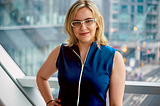 Sarah Kurchak, a white woman with blonde hair and white glasses, smiles into the camera for a portrait. She wears a dark blue shoulderless zip-up top and stands in front of a window that looks out over a city junction.