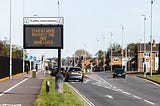 Vehicles on a road in the UK next to an electronic sign, where it reads: “Stay at Home. Protect the NHS. Save lives.”