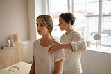 A therapist gently evaluates a seated woman’s shoulder posture during a physical therapy or body alignment session. The setting is calm and bright, with natural light streaming through large windows. Reflecting the perspective of Ramneek Bhogal on the holistic approach to managing tennis elbow.