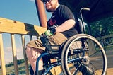A light-skinned Black trans man in a black shirt and tan shorts, sitting in a wheelchair on a deck outside.