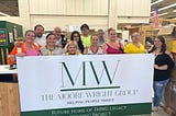 A dozen staff members from The Moore Wright Group pose together in the organization’s warehouse behind a white sign with green font announcing their Legacy Housing.
