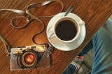 A tabletop photograph of a Leica M6 camera with a Voigtlander 35mm lens and a delicious cup of coffee on the side. The camera has an odd-looking hand-made half-case made from foamy and a brown bandana with white motifs. The photographer is wearing his favorite old blue jeans and Stan Smith Adidas sneakers with red details.