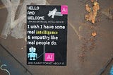 A sticker on a rusted metal background. The background of the sticker is black. The sticker shows a simple illustration of a small blue robot toy, the letters ‘AI’ in a pink square at the top and bottom, and the words, “Hello and welcome. Artificial intelligence. I wish I have some real intelligence and empathy like real people do. And funny? Forget about it.”