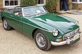 A British racing green MGB open-topped roadster sports car from the 1960s stands on gravel outside a house in England