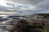 View from the cliffs at Llantwit Major beach at low tide, with rocky tidal flats, dramatic cloudy sky, sunlight on the sea, and a small building by the shore.
