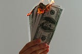 A hand holding a stack of cash that has caught on fire