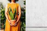 A woman in an earth-tone orange outfit holding a potted plant against a white and concrete gray background with additional plants