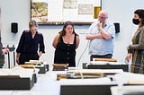 Four people looking at collections material on a table, some fragile books are lying open on book supports other books sit closed in piles.