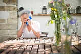 Old woman sitting at a table with one hand covering half her face and the other holding a bright blue phone to her ear. She looks distressed.
