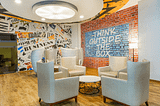 A well lit corner inside an office space with white comfy couches around a white centre table with wall graffiti in the background written “Think Outside the Box”.