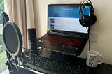 Foto da estação de trabalho, com uma mesa, um notebook suspenso por um suporte, um microfone preso por uma haste, um teclado, um fone de ouvido grande pendurado no notebook, e uma caneca branca escrito Jornalismo.