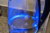 A glass electric kettle on a kitchen counter. The electric kettle glows blue, indicating that it has been turned on. There is condensation visible on the inside of the kettle. The handle is made of black plastic, and the stand is also black. The counter is made of a speckled pattern.