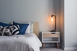 A low bed with a white bedspread and blue accent pillows. Next to it is a simple nightstand with a metal arm lamp with a clear shade, with a lit yellow light bulb inside it. Indirect sunlight comes from off screen to the right, lighting the entire scene.