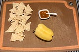 A strip of corn chips, a little bowl of hot sauce with a small ceramic spoon, and a bell pepper on a paper composite serving board