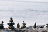 Stacks of balanced stones on a beach next to water. The picture is very calm and serene, in tones of light grey and subtle blue.