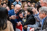 Joe Biden in a blue suit smiles, posing for a selfie with a supporter as a crowd surrounds him