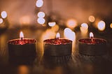 Shows three pink tealight candles burning in the dark with blurry lights behind them