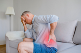 The image shows a man sitting on a couch, with one hand on his head and the other on his lower back. He seems to be in pain, specifically in the lower back region, which is highlighted in red to emphasize discomfort or pain.