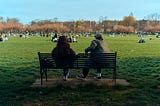This picture shows a man and a woman sitting on a bench. We are looking at them from behind and we can see the park area they are facing that appears to have dozens of people enjoying nature. The man has his arm propped up on the back of the bench and his posture is turned towards the woman, indicating he may be talking to her even though he appears to be looking out away from her. She is facing straight, potentially not engaged, also looking out at the park.
