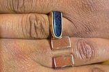 Two brown hands interlock at the third fingers, placing the engagement rings’ blue and copper goldstones parallel to each other.