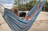 What No One Tells You About Remote Work and Being a Digital Nomad- A women laying in a hammock on the beach holding an ipad.