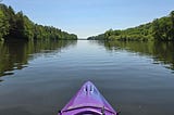 The nose of a purple kayak is centered with a wide lake channel ahead. Low hills covered in green forests are on both sides. The water is calm and there are no buildings, people, or other boats in sight.