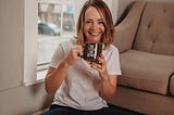 Woman sitting on the floor smiling with a coffee cup reading Be You Be Kind.
