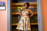 A open closet. Tamarah is standing in front wearing a white floral dress and holding a green Ryobi brad nailer. Behind her are 3 floating shelves she built. The closet wall, closet door and the trim are a blue/dark grey. The room color is pink and there is a stripe of pink in the middle top inside the closet.