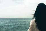 Photo from behind of a woman with long dark hair and a white cotton eyelet looking out at the horizon and the meeting of an aqua colored ocean and pale white skies.