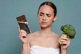 Young woman with acne holding a block of chocolate and broccoli