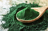 A close-up of a wooden spoon cradling a heap of fine, bright green spirulina powder, with some scattered on the surface beneath it, highlighting the powder’s texture.