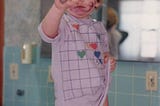 Young, redheaded girl in lavender, geometric patterned nightgown with red, green, yellow and blue hearts. Purple marker ink covers the child’s hands, face and legs.