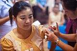 A woman receives a vaccination in her upper arm administered by a healthcare worker. She is seated and looking away, while the healthcare worker carefully holds her arm and prepares the injection. The setting appears to be a clinic or health facility with other people in the background.