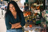 Ivelyse Andino, an Afro-Latina woman with long dark curly hair, stands in front of an open air market, with her arms crossed, smiling, wearing a blue three-quarter sleeve shirt, two silver necklaces, and blue jeans.