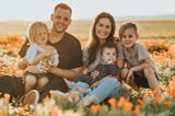 Family, 2 parents and 3 kids, sitting in a sunny field with lots of orange and white flowers