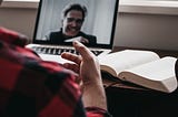 image shows a person’s back and hand from behind as they are looking at another person on the computer screen