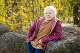 A boy with cancer posing outside by a rock.