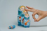 Four blue Russian nesting dolls lined up in a row. The smallest one lays on its side. A hand flicks the second smallest doll